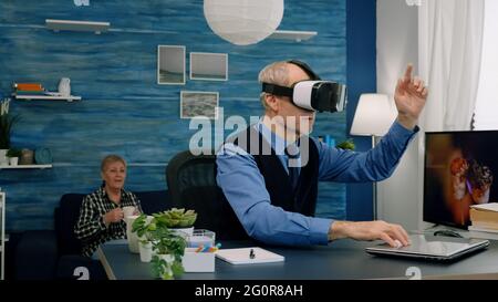 Senior man using virtual reality goggles in living room gesturing sitting in home workplace. Retired man experiencing vr headset while woman watching tv sitting on sofa in background. Stock Photo