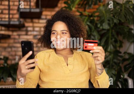 Online payment. Young happy african-American woman holds bank card and smartphone sitting on the armchair at home, making purchase online, ordering food delivery, shopping and paying online Stock Photo