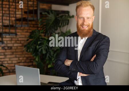Smiling young redhead high skilled leader in formal suit looks at the camera, confident bearded businessman stands in office with arms folded Stock Photo