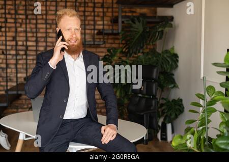 Smiling young redhead high skilled leader talking on the smartphone, ceo has phone conversation standing in modern office, red haired man in formal suit chatting on the cellphone Stock Photo