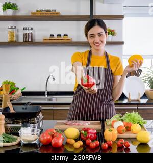 Young housewife stand smiling,  hold red and yellow bell pepper with both hands. Offering the red one on in the front. The kitchen counter full of var Stock Photo