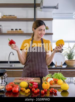 Young housewife stand smiling,  hold red and yellow bell pepper with both hands. Looking at the yellow one on the left. The kitchen counter full of va Stock Photo