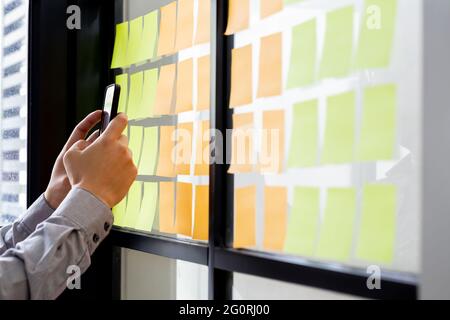 IT worker tracking his tasks on kanban board in mobile phone application. Using task control of agile development methodology. Man attaching sticky no Stock Photo