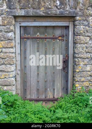 A old wooden door in a stone wall, with rusting hinges and metal studs and weeds in foreground. Stock Photo