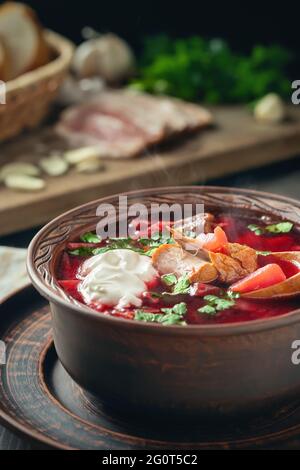 Freshly cooked borscht - traditional dish of Russian and Ukrainian cuisine in earthenware dishes with bacon, sour cream and garlic, vertical image. Stock Photo