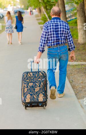 A man drags a suitcase on wheels along the sidewalk in the city. Back view Stock Photo