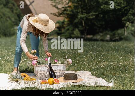 Unrecognizable young woman with straw hat arranging flowers into a basket. Female setting a blanket for picnic with wine bottle and variety of fruits. Stock Photo