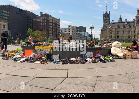 Ottawa, Canada - June 2, 2021: People gather round and leave shoes and toys left near the Centennial Flame on Parliament Hill in memory of the 215 children whose remains were found near of former Residential School in Kamloops, B.C. There is a sign referring a current lawsuit the Assembly of First Nations has against the Canadian government. Stock Photo