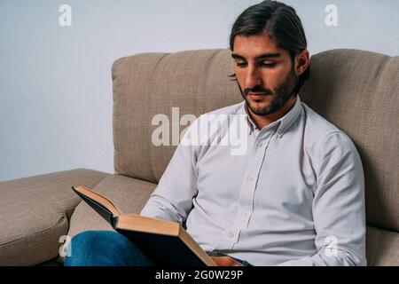 Elegant Hispanic Man with Long Hair and Bear Sitting Concentrated at Couch Reading Holy Bible Stock Photo