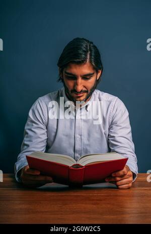 Hispanic Elegant Good Man with Beard Reading Red Book Sitting on a Wooden Table Concentrared Study Stock Photo