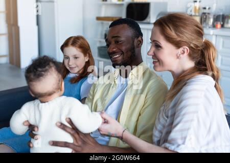 happy interracial family looking at infant baby girl while sitting at home Stock Photo