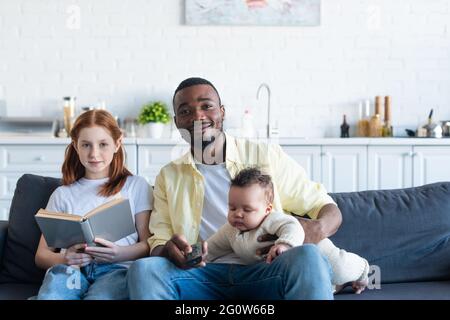 preteen girl holding book near african american father with infant sister watching tv at home Stock Photo