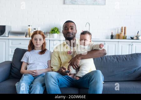 happy african american man holding infant baby while watching tv near preteen daughter Stock Photo
