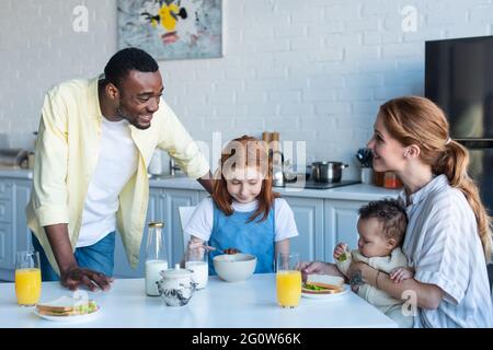 happy african american man standing near multiethnic daughters and wife having breakfast in kitchen Stock Photo