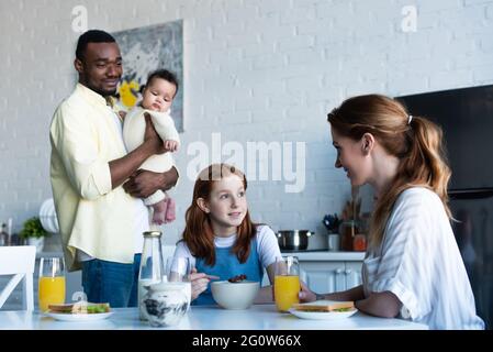 african american man holding infant near wife with daughter having breakfast in kitchen Stock Photo