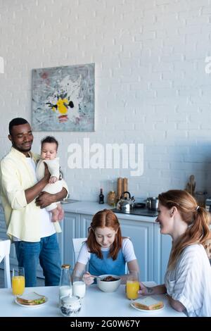 happy african american man holding infant daughter near family having breakfast in kitchen Stock Photo