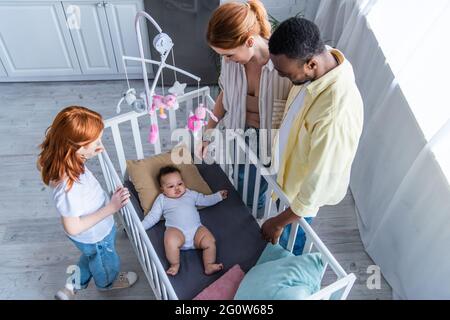 overhead view of multiethnic family looking at infant girl lying in crib Stock Photo