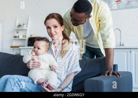 african american man looking at happy wife sitting on sofa with infant daughter Stock Photo