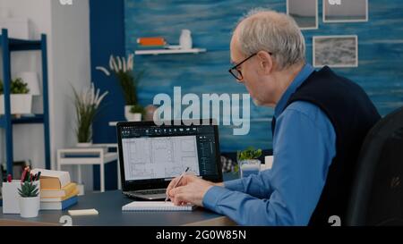 Senior man architect analysing digital prototype with plans from laptop working from home, taking notes on notebook. Elderly engineer designer using cad software to create a 3D concept of buildings. Stock Photo