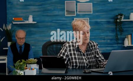 Multitasking retired woman reading on laptop and tablet in same time working from home. Focused old remote employee searching, analysing financial reports while elderly husband reading a book Stock Photo