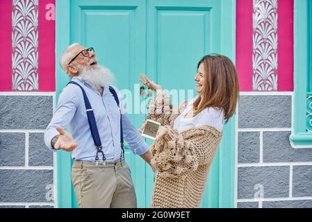 Happy middle aged woman sharing data in social media with stylish senior man while browsing mobile phone on street against colorful building Stock Photo