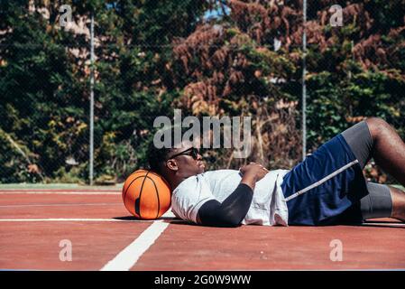 Black Afro guy lying on his ball on the basketball court while using his cell phone. Concept of technology and sport. Stock Photo