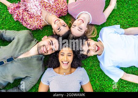 Group of five happy young diverse multiracial gen z friends lying in circle head to head on the grass of a park lawn looking to the camera from the bo Stock Photo