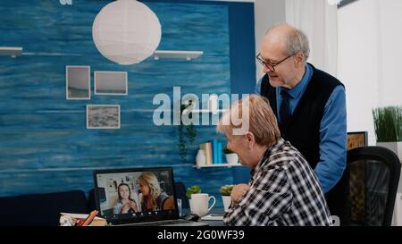 Excited senior woman and her husband having video conference on laptop, speaking with remote nephews sitting in living room. Online call with daughter and niece using modern communication online Stock Photo
