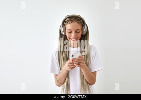 Positive female with long braids wearing white t shirt standing against white background and enjoying songs in headphones while using mobile phone Stock Photo