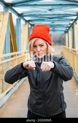 Fit sporty middle aged blonde 40s woman in red cap doing jogging workout, stretching, enjoying fitness and healthy lifestyle at city street at spring Stock Photo