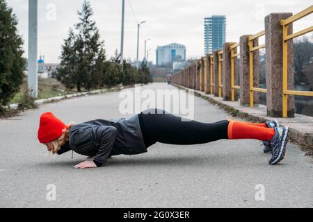 Fit sporty middle aged blonde 40s woman in red cap Standing in Plank, doing workout, stretching, enjoying fitness at city street at spring autumn day. Stock Photo