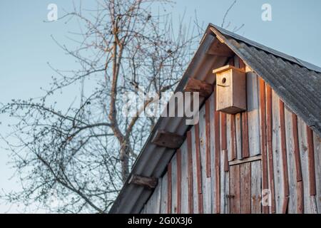Birdhouse hanging under roof of old wooden shed next to tree against blue sky. House for birds Stock Photo