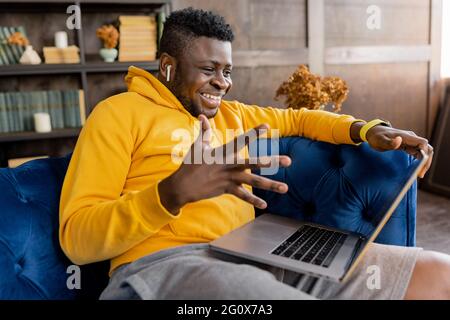 Joyful black businessman sitting on sofa at home, looking at the computer screen, emotionally talking with friend, colleagues. African American worker Stock Photo