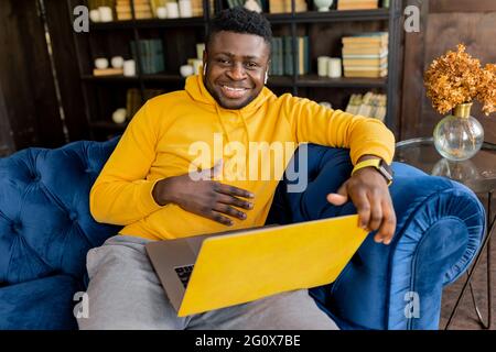 African American man sitting on sofa sincerely smiling looking at camera laughing,joyful male student freelancer having online lesson, meeting on Stock Photo