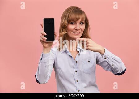 A middle-aged girl with shows on the phone screen, the blonde holds the phone with a screen on a pink background Stock Photo
