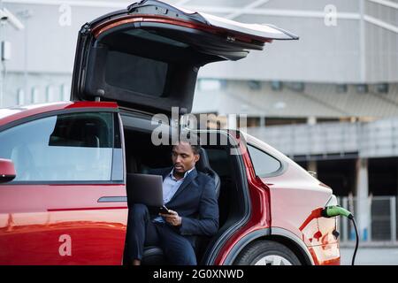 African american businessman in stylish suit using modern laptop for remote work while his luxury electric car is charging. Handsome male sitting in side auto with opened door and looking aside. Stock Photo