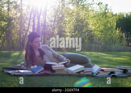 Teen girl, young woman relaxing on blanket outside reading a book surrounded by piles of books in summer with sunset behind her Stock Photo