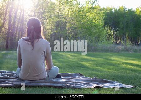 Young woman sitting on blanket facing away from camera outdoors on green grass with trees and sun setting in background, with copy space Stock Photo