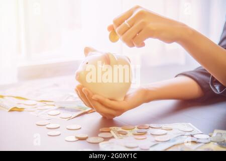 Coins on hand are dropped Piggy bank with cash pile on wooden top table, money savings concept. Stock Photo