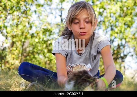 Defocus blonde little expression girl playing and caress cat, black and white small kitten. Nature blurred green summer background. Girl stroking pet. Stock Photo