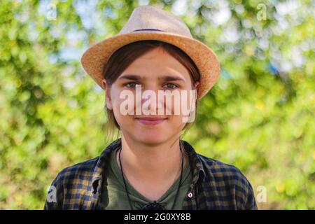 Defocused close-up light smiling young brunette caucasian woman in hat. Summer blurred green nature bokeh background. Portrait of handsome young cute Stock Photo