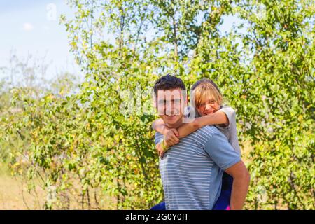 Defocused brother giving sister ride on back. Portrait of happy girl on man shoulders, piggyback. Family playing outside. Summer holidays smiling peop Stock Photo