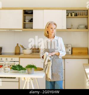 Beautiful senior woman in kitchen apron wiping hands during cooking, looking in window with smile and enjoying life after retirement. Smiling retired female preparing healthy food for family at home Stock Photo