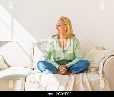 Enjoying life after retirement. Happy senior woman looking in window and smiling, expressing happiness while sitting on sofa and relaxing at home. Cheerful retired female spending time in living room Stock Photo