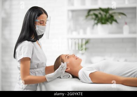 Side view of brunette beautician in protective mask doing beauty injection for improvements skin face. Concept of procedures for tighten and moisturize face skin. Stock Photo