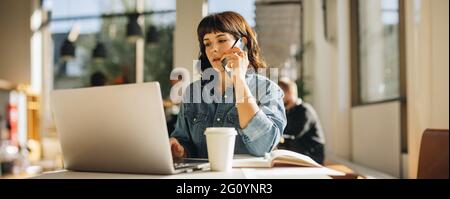 Young female sitting at office desk talking on mobile phone and working on laptop. businesswoman talking on smart phone while using laptop in coworkin Stock Photo