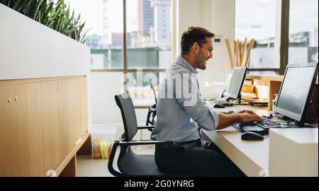 Manager smiling while working on computer. Happy young man sitting in office coding on computer. Stock Photo