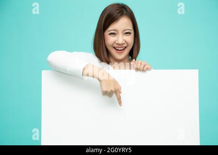 Smiling happy Asian woman standing behind big white poster and pointing finger down to blank copy space isolated on light green background Stock Photo