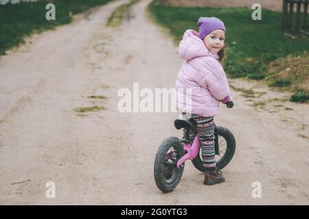Child in pink jacket and lilac hat rides balance bike on dirt road in countryside Stock Photo