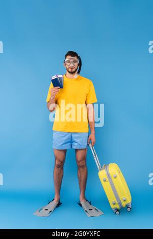 Man in swimming flippers and goggles holding passports and suitcase on blue background Stock Photo